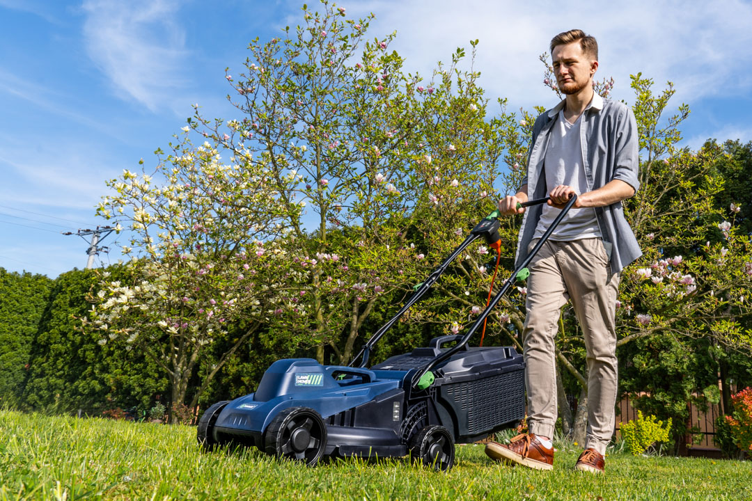 Mężczyzna prowadzi kosiarkę elektryczną po trawie w ogrodzie, w tle drzewa i kwiaty.